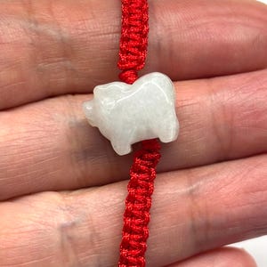 May include: A bracelet featuring a white, translucent, carved stone pig charm centred on a vibrant red braided cord. The cord is constructed with a series of intricate knots, providing a textured appearance that contrasts with the smooth, polished surface of the stone animal figure.