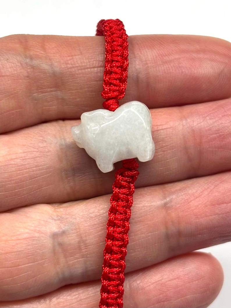May include: A bracelet featuring a white, translucent, carved stone pig charm centred on a vibrant red braided cord. The cord is constructed with a series of intricate knots, providing a textured appearance that contrasts with the smooth, polished surface of the stone animal figure.