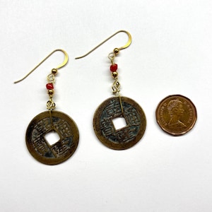 May include: A pair of antique coin earrings with gold-colored hooks and red beads. The earrings feature round, bronze-colored coins with square holes in the center. A penny is included for size comparison.