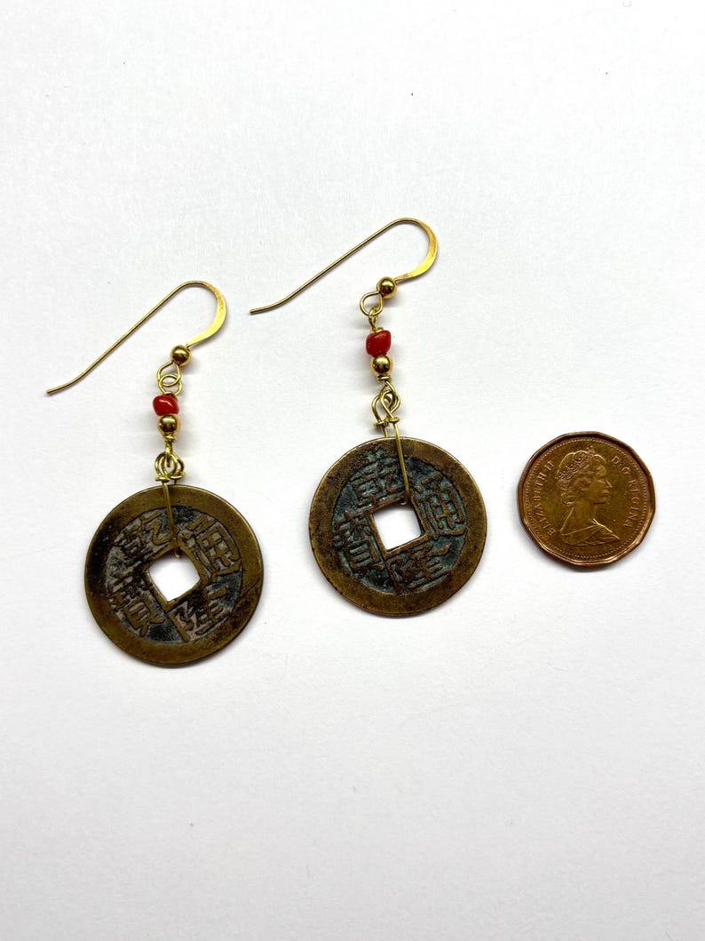 May include: A pair of antique coin earrings with gold-colored hooks and red beads. The earrings feature round, bronze-colored coins with square holes in the center. A penny is included for size comparison.