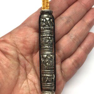 May include: A silver prayer wheel with intricate carvings and black and yellow cords. The prayer wheel is held in a hand.
