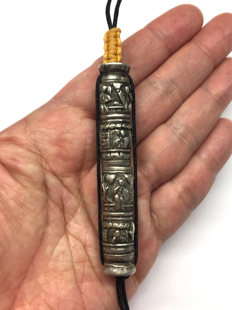 May include: A silver prayer wheel with intricate carvings and black and yellow cords. The prayer wheel is held in a hand.