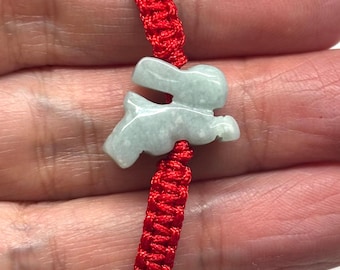 May include: A bracelet featuring a light green, translucent stone carved into the shape of a rabbit. The stone charm is centered on a vibrant red woven cord, which includes an adjustable sliding knot mechanism for a secure fit.