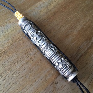 May include: A silver prayer scroll with intricate carvings and a black cord. The scroll is decorated with religious symbols and figures.