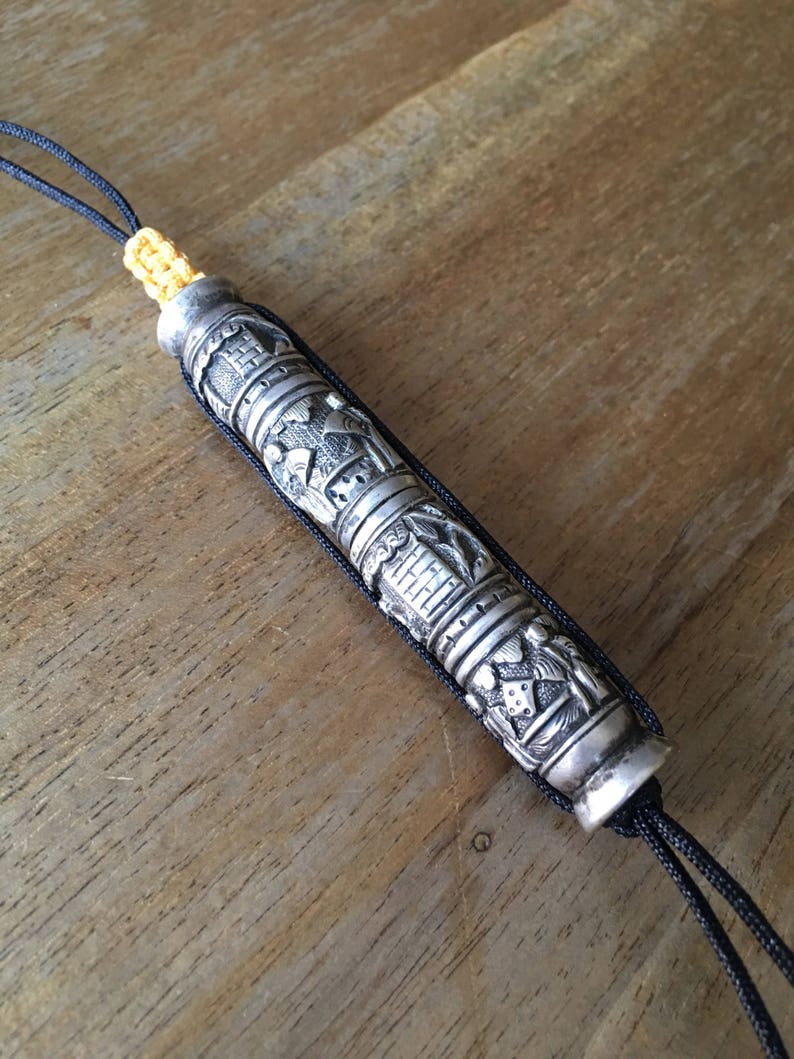 May include: A silver prayer scroll with intricate carvings and a black cord. The scroll is decorated with religious symbols and figures.