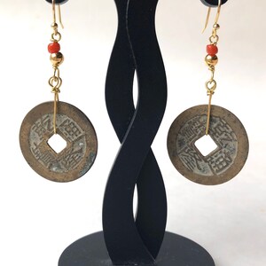 May include: A pair of antique coin earrings. Each earring features a circular, bronze-colored coin with a square center hole, suspended from a gold-colored hook. The earrings include gold-colored beads and small red beads. The earrings are displayed on a black stand.