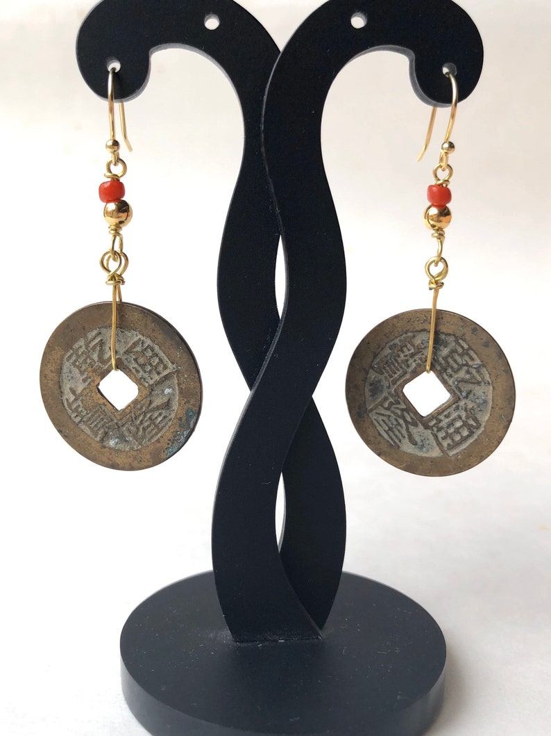 May include: A pair of antique coin earrings. Each earring features a circular, bronze-colored coin with a square center hole, suspended from a gold-colored hook. The earrings include gold-colored beads and small red beads. The earrings are displayed on a black stand.