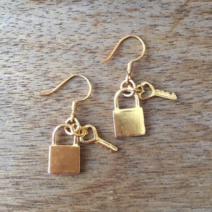 May include: A pair of gold earrings featuring a small padlock and key charm. The earrings are on a wooden surface.