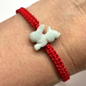 May include: A red woven cord bracelet featuring a central pale green carved stone rabbit charm. The bracelet is constructed from a series of knotted red threads that form a continuous band, with the rabbit charm positioned in the center.