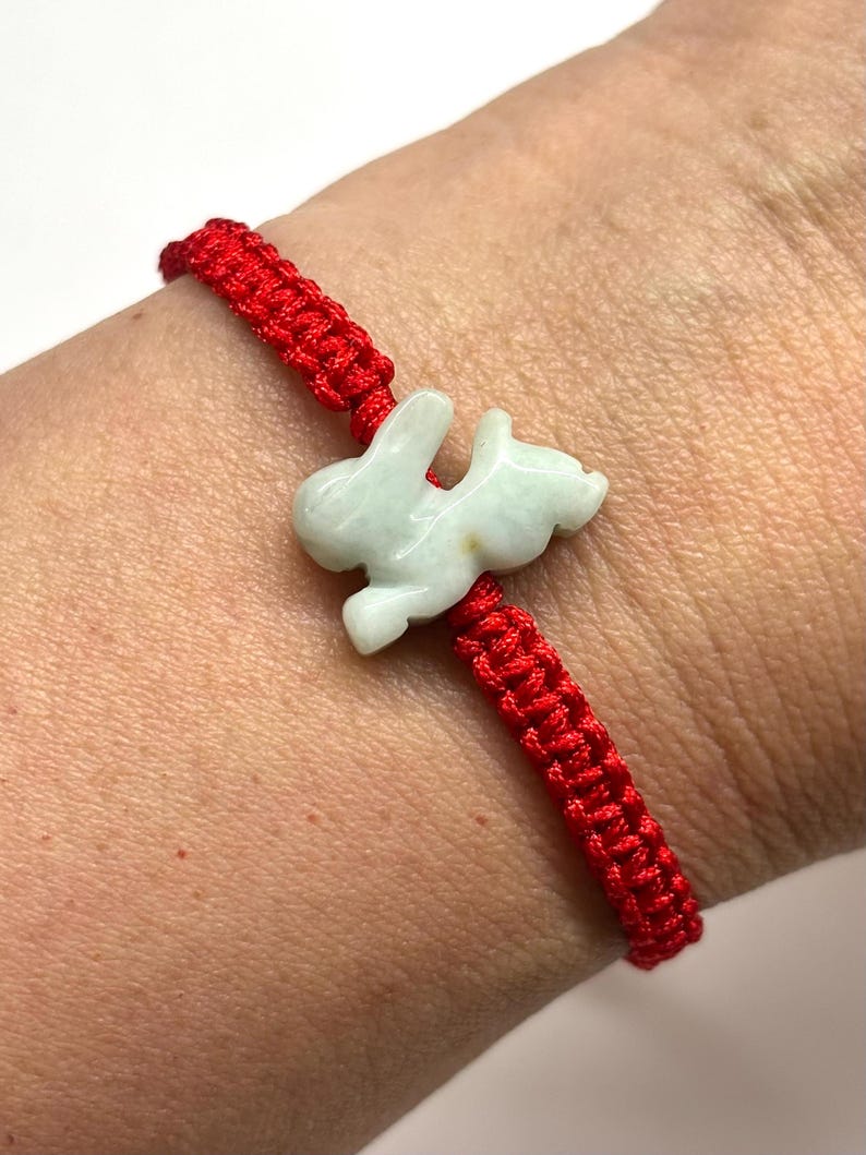 May include: A red woven cord bracelet featuring a central pale green carved stone rabbit charm. The bracelet is constructed from a series of knotted red threads that form a continuous band, with the rabbit charm positioned in the center.