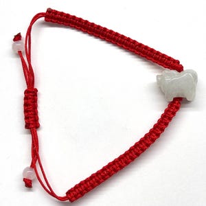 May include: A red cord bracelet featuring a central carved white jade-like animal charm. The bracelet is constructed with a woven macrame-style band and includes an adjustable sliding knot closure finished with two small, round, pale pink beads.