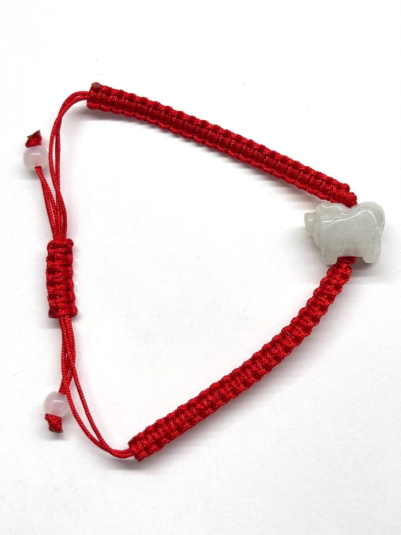 May include: A red cord bracelet featuring a central carved white jade-like animal charm. The bracelet is constructed with a woven macrame-style band and includes an adjustable sliding knot closure finished with two small, round, pale pink beads.