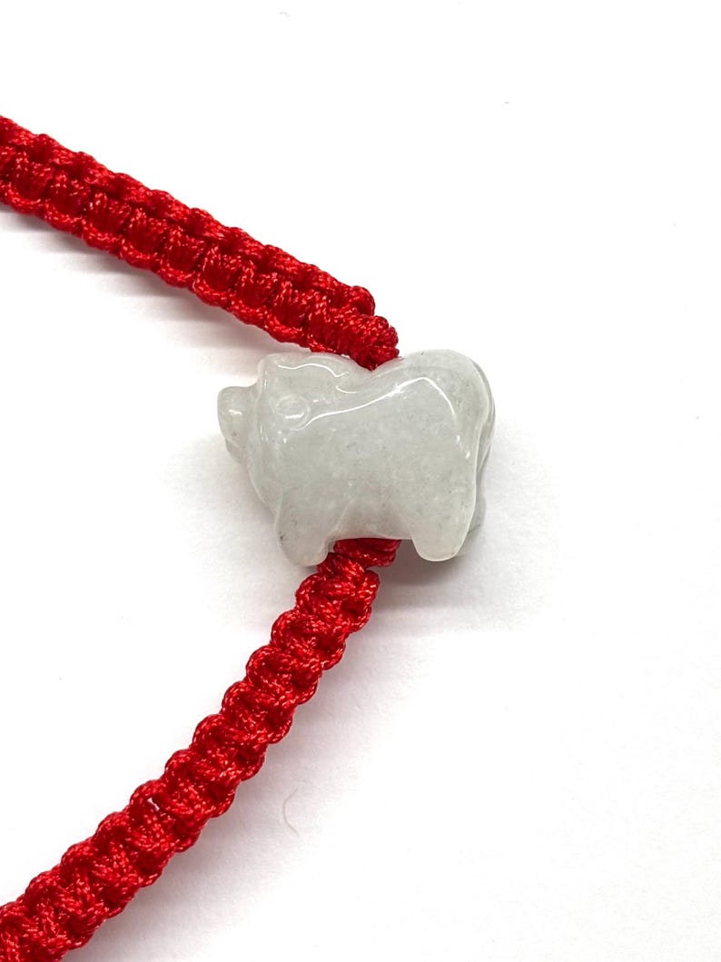 May include: A bracelet featuring a bright red woven cord and a single, carved, pale white stone charm in the shape of a pig. The cord is constructed with a series of interlocking knots, and the stone charm is smooth with a polished finish.