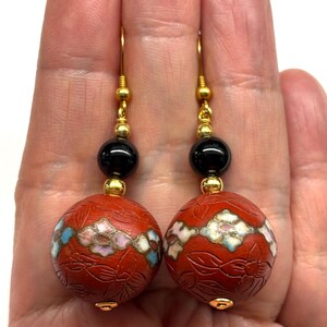 May include: A pair of red and black dangle earrings with gold accents. The earrings feature a round, red bead with a floral design and a black bead with a gold accent.