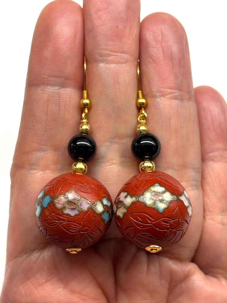 May include: A pair of red and black dangle earrings with gold accents. The earrings feature a round, red bead with a floral design and a black bead with a gold accent.