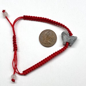 May include: A red cord bracelet featuring a gray carved stone ram charm and two small translucent beads at the adjustable ends. A bronze-colored coin with the profile of Queen Elizabeth II is placed in the center for scale reference.