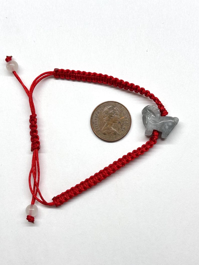 May include: A red cord bracelet featuring a gray carved stone ram charm and two small translucent beads at the adjustable ends. A bronze-colored coin with the profile of Queen Elizabeth II is placed in the center for scale reference.