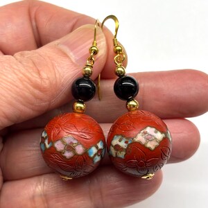 May include: A pair of dangle earrings with red, white, and blue floral designs on the round beads. The beads are attached to black beads and gold hooks.