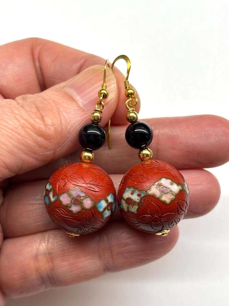 May include: A pair of dangle earrings with red, white, and blue floral designs on the round beads. The beads are attached to black beads and gold hooks.