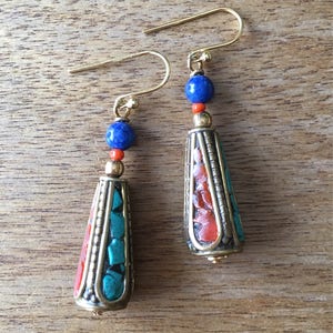 May include: A pair of dangle earrings with a gold tone finish. The earrings feature a teardrop-shaped pendant with a mix of turquoise, red, and orange stones. Each earring is topped with a blue lapis lazuli bead.