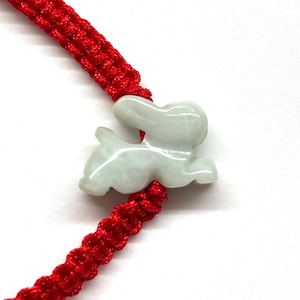 May include: A bracelet featuring a pale green, translucent stone carved into the shape of a rabbit. The rabbit charm is centered on a vibrant red woven cord, which is constructed with a series of intricate, knotted patterns along the band.