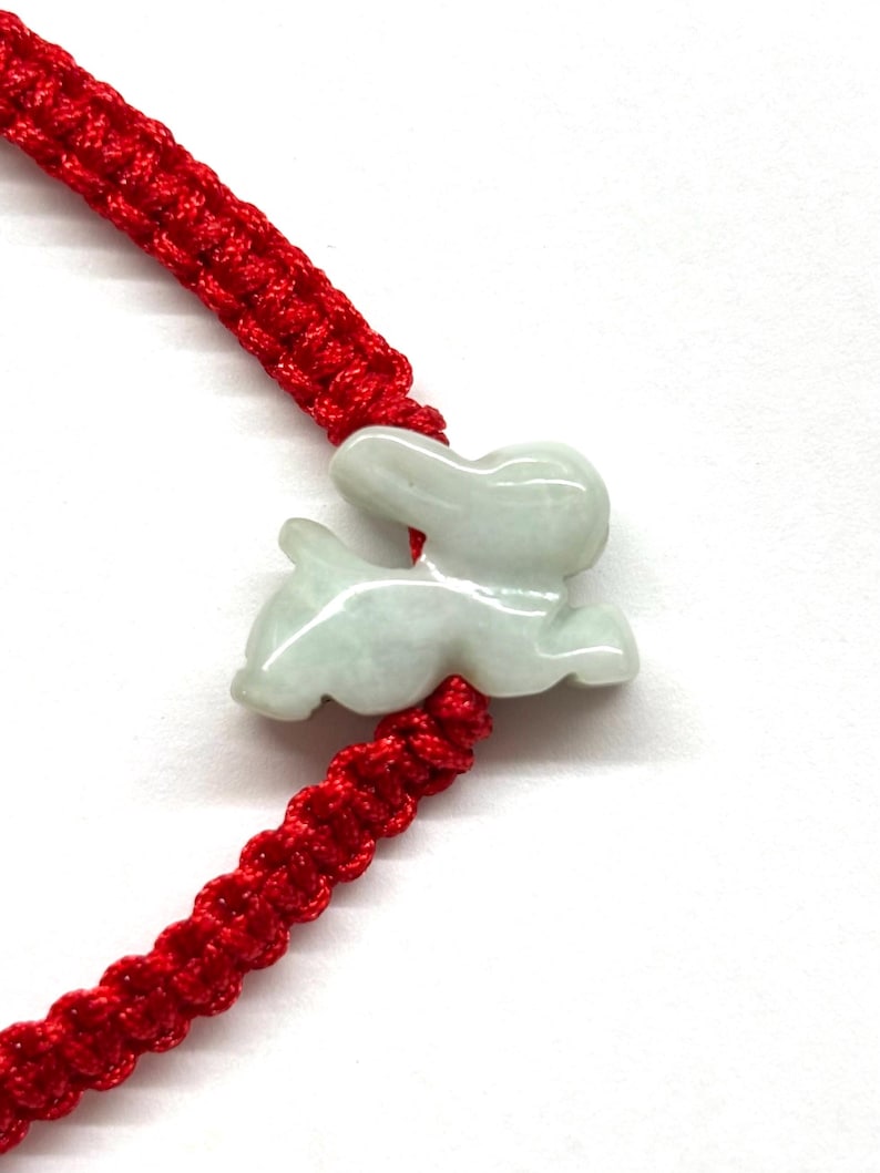 May include: A bracelet featuring a pale green, translucent stone carved into the shape of a rabbit. The rabbit charm is centered on a vibrant red woven cord, which is constructed with a series of intricate, knotted patterns along the band.
