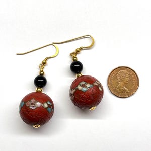 May include: A pair of gold earrings with red and black beads. The red beads are decorated with floral patterns and the black beads are round. The earrings are hanging from gold hooks.