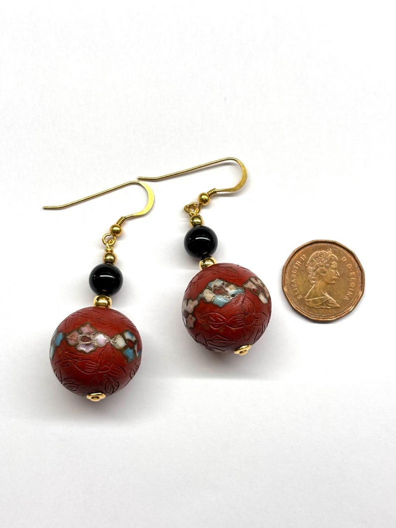May include: A pair of gold earrings with red and black beads. The red beads are decorated with floral patterns and the black beads are round. The earrings are hanging from gold hooks.