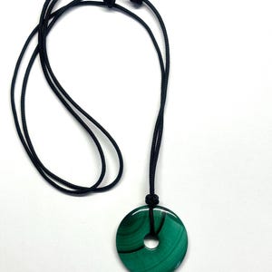 May include: A green malachite stone pendant with a hole in the center, hanging from a black cord necklace.