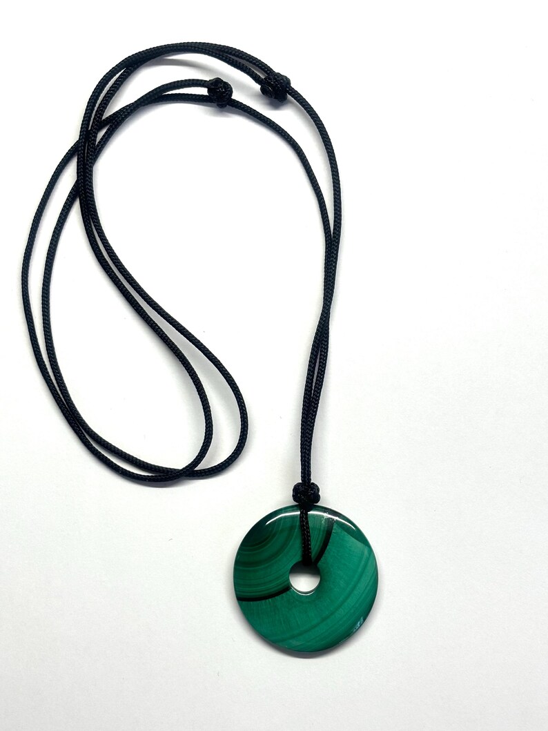 May include: A green malachite stone pendant with a hole in the center, hanging from a black cord necklace.