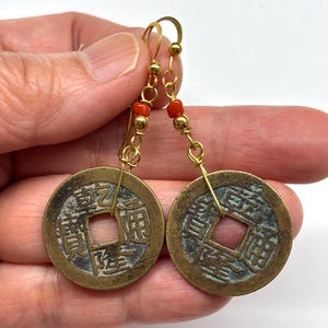 May include: A pair of gold-colored earrings featuring antique Chinese coins with square center holes. The earrings have gold-colored hooks and are adorned with small red beads.