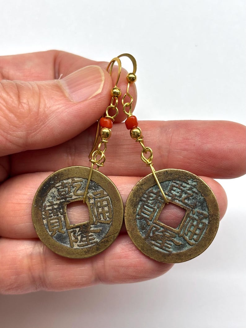May include: A pair of gold-colored earrings featuring antique Chinese coins with square center holes. The earrings have gold-colored hooks and are adorned with small red beads.