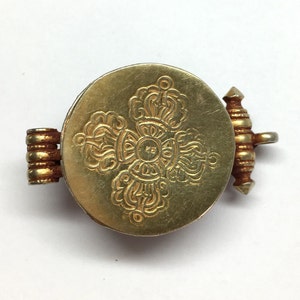 K&ouml;nnte beinhalten: Ein goldfarbener Metallanh&auml;nger mit einem runden Design, das ein kompliziertes, graviertes Muster aufweist. Der Anh&auml;nger hat einen Scharnierverschluss.