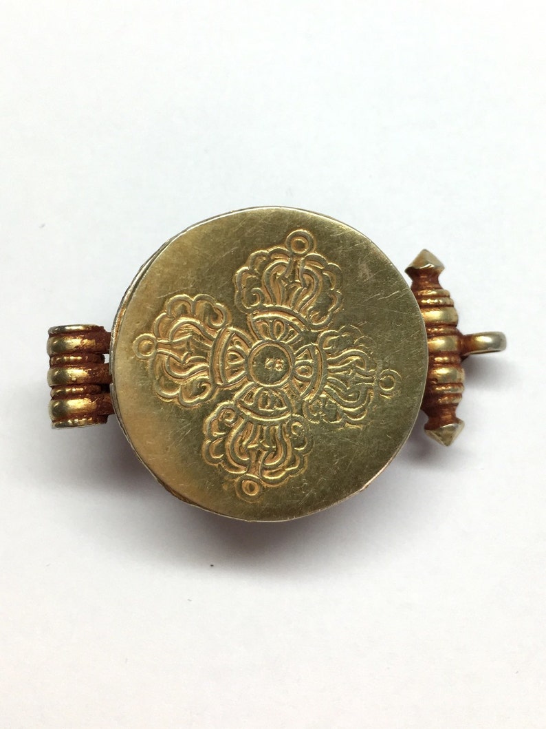 K&ouml;nnte beinhalten: Ein goldfarbener Metallanh&auml;nger mit einem runden Design, das ein kompliziertes, graviertes Muster aufweist. Der Anh&auml;nger hat einen Scharnierverschluss.