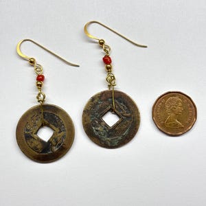May include: A pair of handcrafted earrings featuring antique coin pendants with square center holes. The earrings have gold-colored hooks and are accented with small red beads. A Canadian penny is next to the earrings.