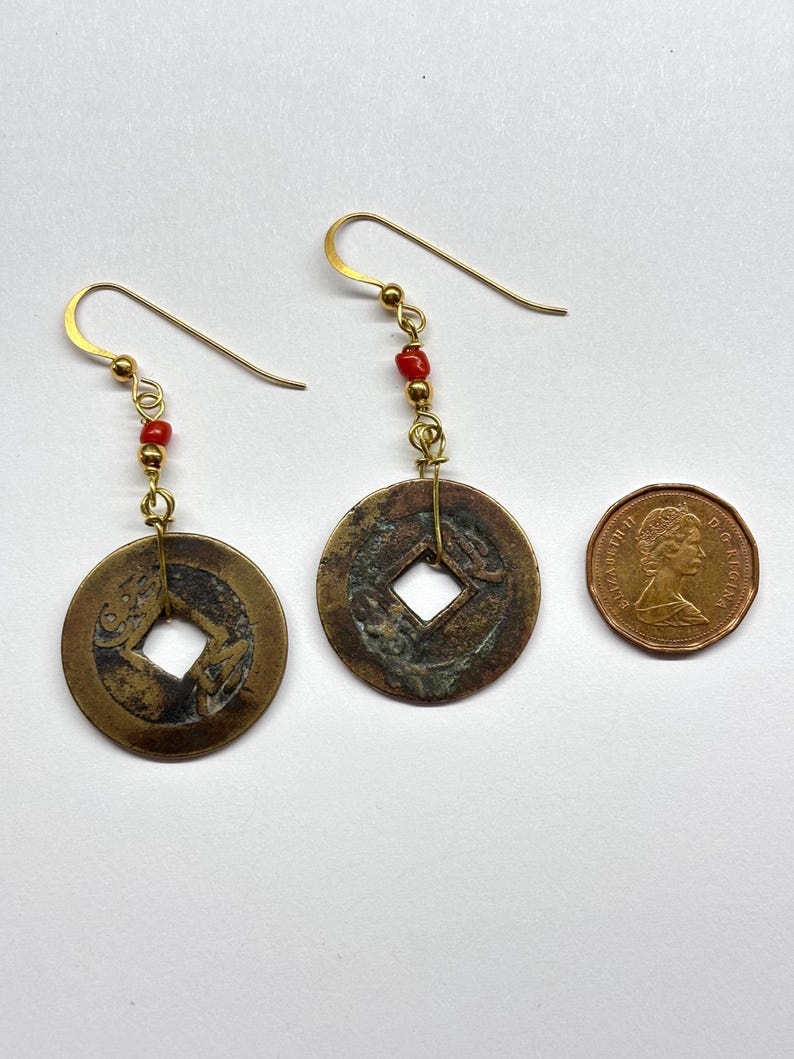 May include: A pair of handcrafted earrings featuring antique coin pendants with square center holes. The earrings have gold-colored hooks and are accented with small red beads. A Canadian penny is next to the earrings.