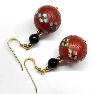 May include: A pair of dangle earrings with red and black beads. The red beads are round and have a floral design. The black beads are round and smooth. The earrings are made of gold metal.
