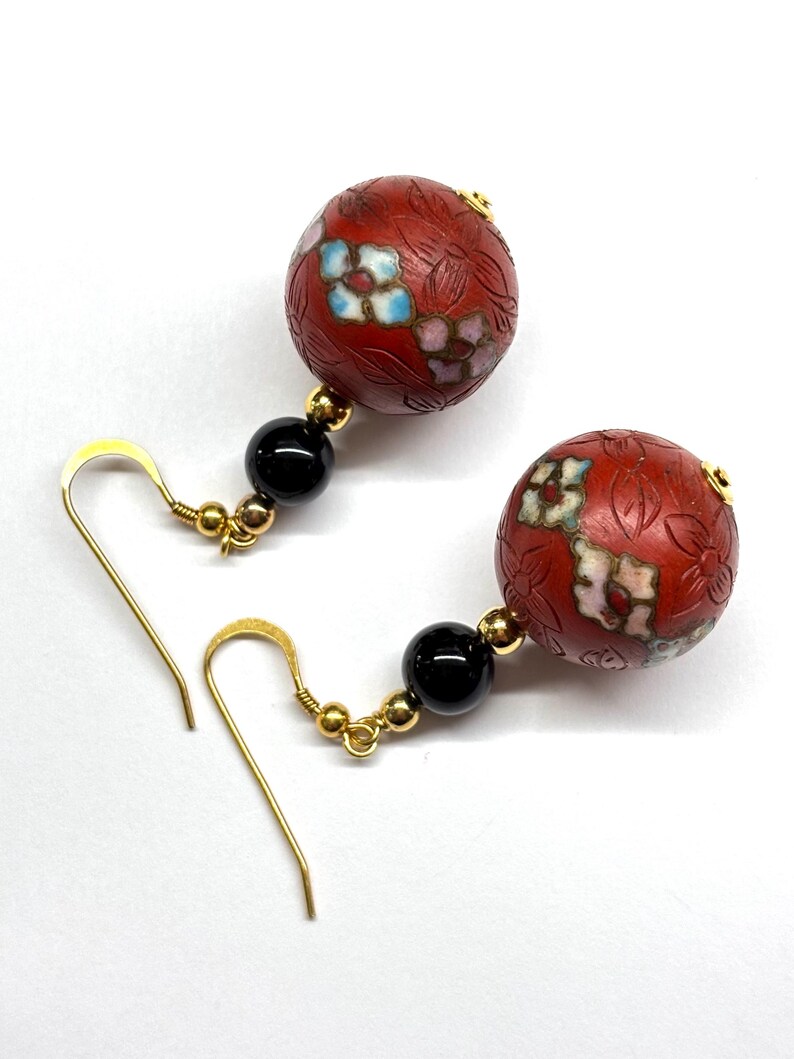 May include: A pair of dangle earrings with red and black beads. The red beads are round and have a floral design. The black beads are round and smooth. The earrings are made of gold metal.