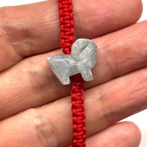 May include: A bracelet featuring a bright red woven cord and a central light gray stone charm carved into the shape of a ram. The smooth, polished stone figure displays the distinct curved horns and body of a ram, securely threaded onto the vibrant red string.