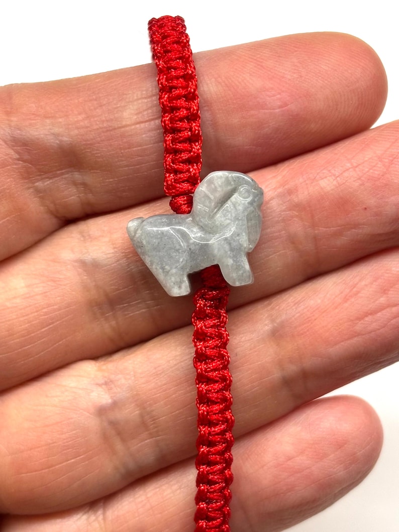 May include: A bracelet featuring a bright red woven cord and a central light gray stone charm carved into the shape of a ram. The smooth, polished stone figure displays the distinct curved horns and body of a ram, securely threaded onto the vibrant red string.