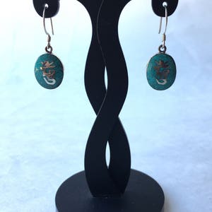 May include: A pair of silver earrings with turquoise stones and a Chinese character etched into each stone. The earrings are hanging from a black earring stand.