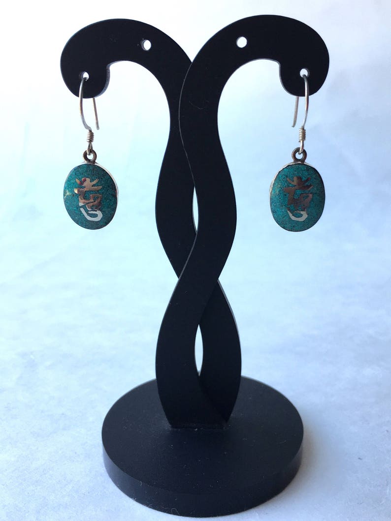 May include: A pair of silver earrings with turquoise stones and a Chinese character etched into each stone. The earrings are hanging from a black earring stand.