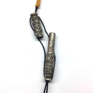 May include: Two silver prayer wheels with intricate designs, connected by a black cord with a yellow tassel. The wheels are cylindrical and have a textured surface.