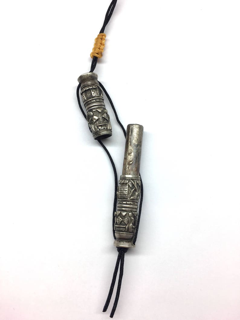 May include: Two silver prayer wheels with intricate designs, connected by a black cord with a yellow tassel. The wheels are cylindrical and have a textured surface.