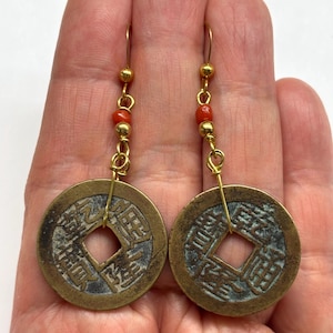 May include: A pair of gold-toned earrings featuring ancient Chinese coins with square center holes. Each earring has a series of small red beads and gold-toned accents. The coins have detailed inscriptions and a weathered appearance.