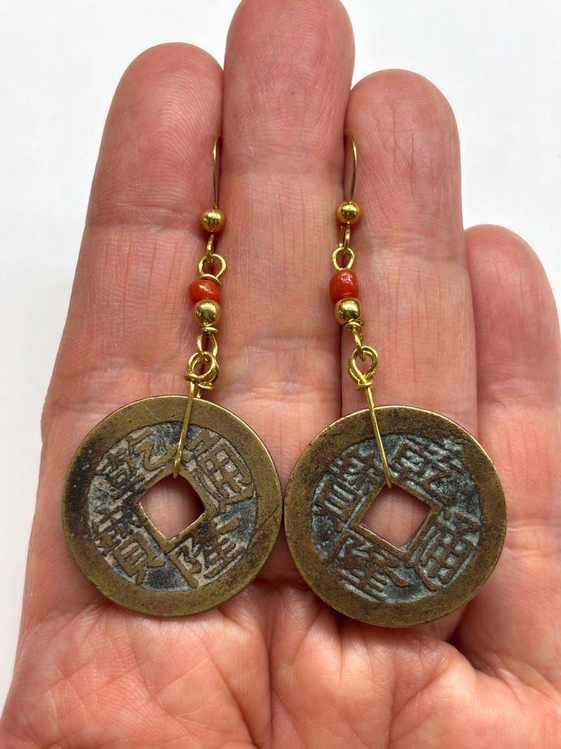 May include: A pair of gold-toned earrings featuring ancient Chinese coins with square center holes. Each earring has a series of small red beads and gold-toned accents. The coins have detailed inscriptions and a weathered appearance.