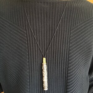 May include: A silver pendant necklace with a cylindrical charm featuring intricate engraved designs. The charm is suspended from a black cord with a yellow bead at the top.