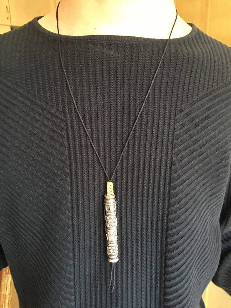May include: A silver pendant necklace with a cylindrical charm featuring intricate engraved designs. The charm is suspended from a black cord with a yellow bead at the top.