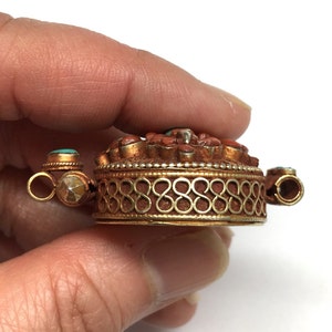 K&ouml;nnte beinhalten: Ein goldfarbener Anh&auml;nger mit einem runden Design, das mit roter Koralle und t&uuml;rkisfarbenen Steinen besetzt ist. Der Anh&auml;nger hat einen dekorativen Rand und zwei &Ouml;sen zum Anbringen einer Kette.