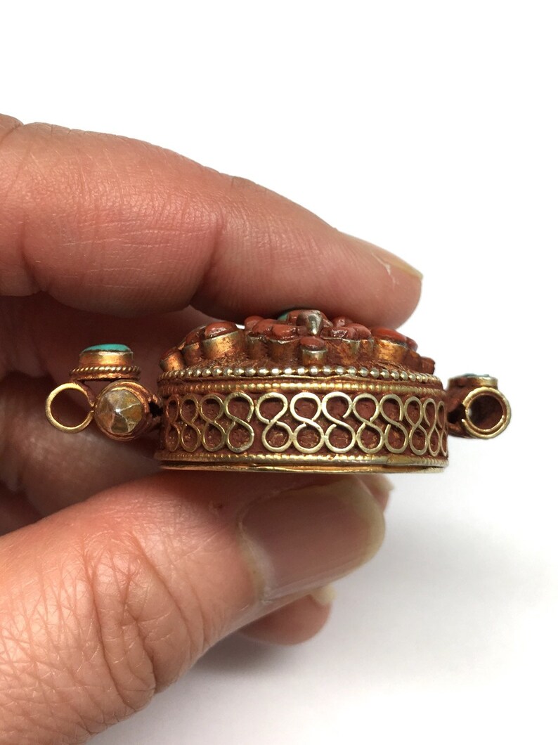 K&ouml;nnte beinhalten: Ein goldfarbener Anh&auml;nger mit einem runden Design, das mit roter Koralle und t&uuml;rkisfarbenen Steinen besetzt ist. Der Anh&auml;nger hat einen dekorativen Rand und zwei &Ouml;sen zum Anbringen einer Kette.